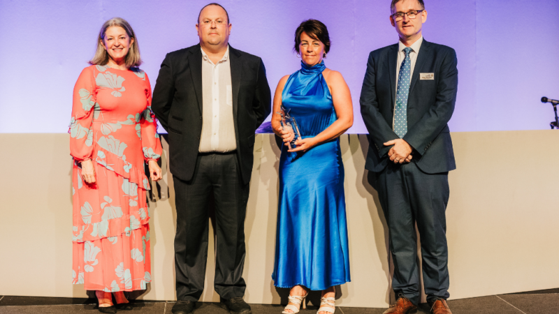 Four people on stage, Rhonda is in the middle holding a glass award