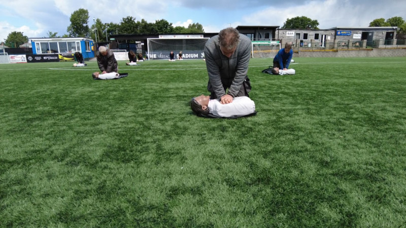 People practicing CPR on manikins on a grassy football field, under a cloudy sky.