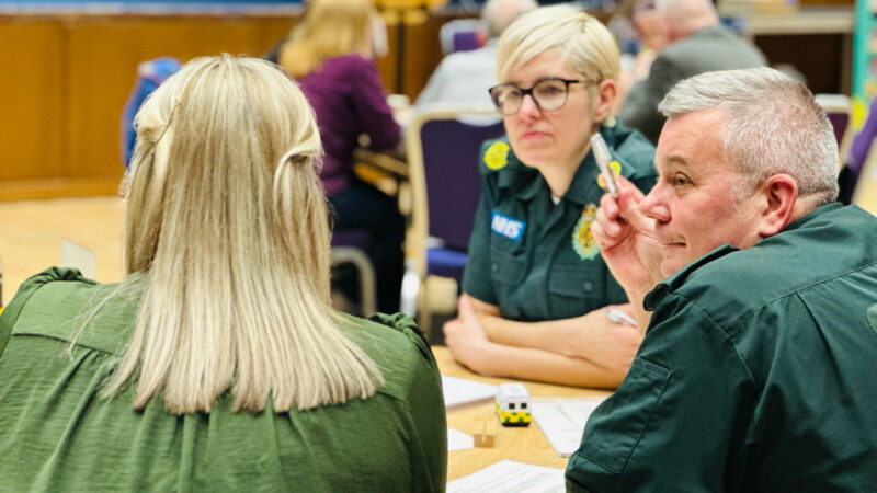 Three people sat round a table talking a woman with her back to the camera has long blonde hair in the middle is a paramedic with short blonde hair and on the right talking to the other woman is a male paramedic with short grey hair.