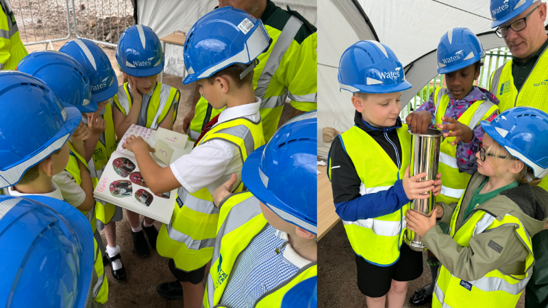Two groups of children in safety vests and helmets gather around each other, one group is holding some artwork, and the other is holding a long silver time capsule.