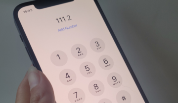 A person holding a smartphone with a dial pad displayed on the screen, showing the digits from 1 to 9, an asterisk, and hash symbol. The phone displays "Add Number" beneath a dialled number of '111 2' at the top, with the time 15:43.