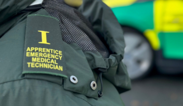 Close-up of a jacket with an "Apprentice Emergency Medical Technician" epaulette, with an ambulance blurred in the background.