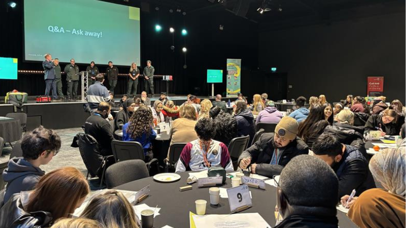 Audience members sit at round tables listening to speakers during a Q&A session at a conference event.